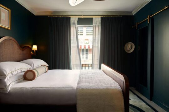 Luxurious dark teal bedroom featuring a king-size bed with a rattan headboard, crisp white linens, and decorative pillows. A large window with dark drapes offers a view of a cityscape, and a sliding barn door with a mirror is visible.