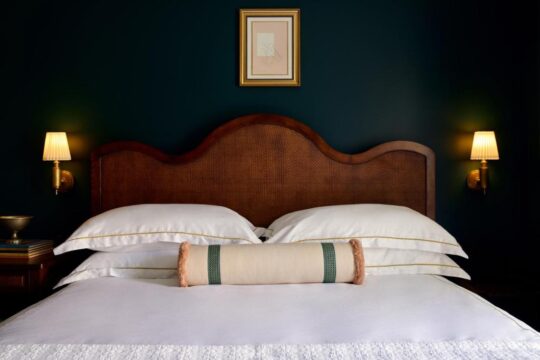 Luxurious hotel bedroom with a plush king-size bed, crisp white linens, a woven rattan headboard, and warm ambient lighting from bedside sconces.