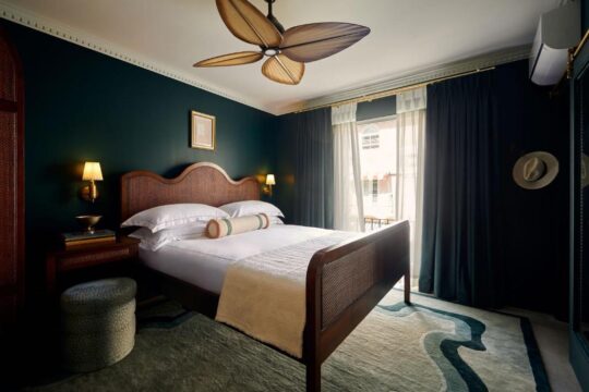 A luxurious hotel bedroom features a dark teal wall, a king-sized bed with crisp white linens, and a plush rug. A ceiling fan with wooden blades is visible overhead, and a window with dark curtains is in the background.