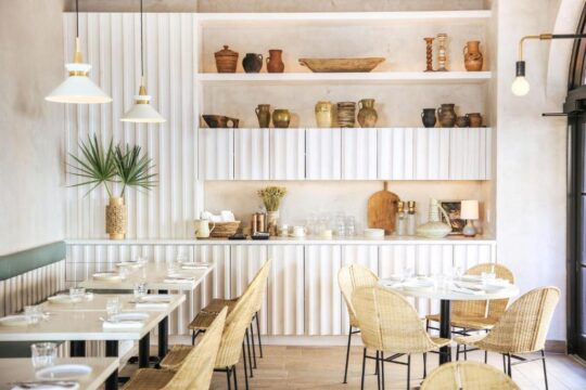 Bright, modern restaurant dining area with white tables, wicker chairs, and decorative shelves filled with pottery and plants.