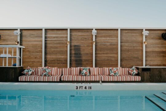 A striped outdoor sofa with floral pillows sits beside a swimming pool, with a slatted wooden wall and speakers in the background. Pool depth marker reads "3 FT 4 IN".