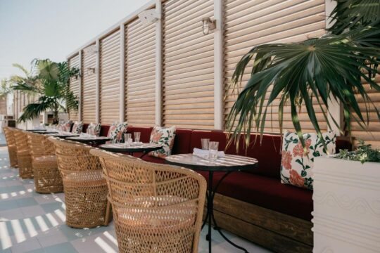 Rattan chairs and a deep red banquette with floral pillows create a chic outdoor dining scene, with slatted wooden walls and palm fronds suggesting a stylish patio restaurant.