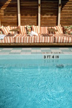 A poolside lounge with striped orange and white cushions and a white rolled towel sits next to a bright blue swimming pool with a depth marker reading "3 FT 4 IN".