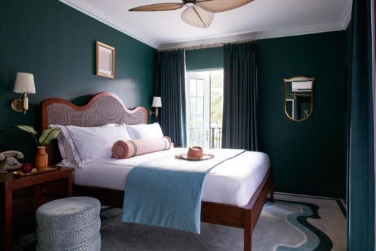 Luxurious hotel bedroom with a king-size bed, dark green walls, and a ceiling fan. A plush blue throw and decorative pillows adorn the bed, with a vintage rotary phone on the bedside table.