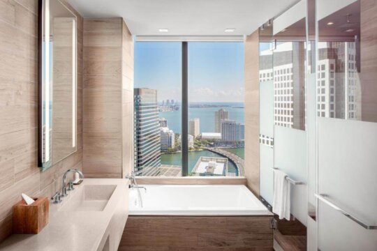 Luxury hotel bathroom with a deep soaking tub positioned before a floor-to-ceiling window offering panoramic city and ocean views, showcasing modern design and upscale amenities.
