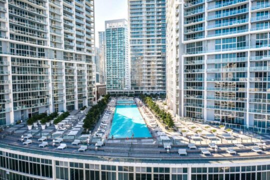 Aerial view of a resort-style swimming pool surrounded by lush greenery and modern high-rise apartment buildings, with lounge chairs and umbrellas set up for relaxation.