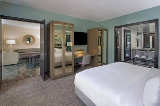 Luxurious hotel suite with a king-size bed, mirrored wardrobes, and an adjoining living area with a sofa and armchair. The room features teal walls and elegant decor.