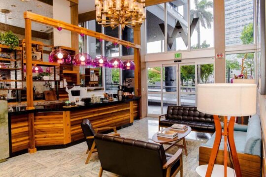 Modern cafe interior with a wooden counter, purple pendant lights, and comfortable seating. An "EXIT" sign is visible in the background.