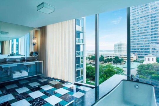 Luxury hotel bathroom with a large soaking tub and double vanity, featuring a stunning panoramic city and ocean view through floor-to-ceiling windows. Black, white, and teal marble floor.