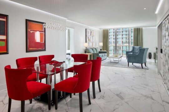 Modern dining room with a glass-top table and six red chairs, adjacent to a living area with a blue sofa and armchair, featuring large windows with city views.