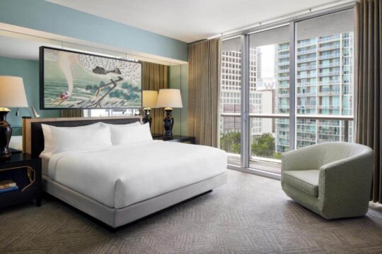 Luxurious hotel bedroom with a king-size bed, modern armchair, and large window overlooking a city skyline. The room features a contemporary art piece and ambient lighting.