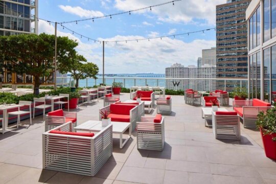 Rooftop terrace lounge with white modern outdoor furniture with red cushions, set against a backdrop of Miami skyscrapers and a clear blue sky.