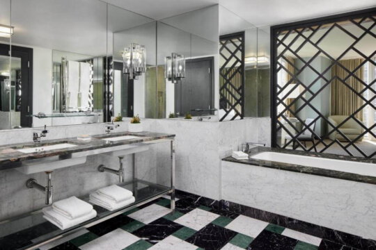 Luxurious modern bathroom with a marble double vanity, geometric patterned screen, and a freestanding bathtub. The floor features a bold black, white, and green diamond tile pattern.
