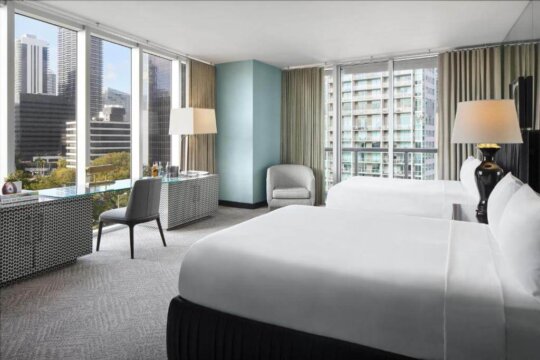 Modern hotel room with two queen beds, floor-to-ceiling windows offering city views, a desk with a chair, and a comfortable armchair.