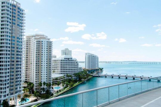 Panoramic view of a modern cityscape with tall, white high-rise buildings overlooking a vibrant blue bay. A bridge spans the water, connecting to distant structures under a bright, partly cloudy sky.