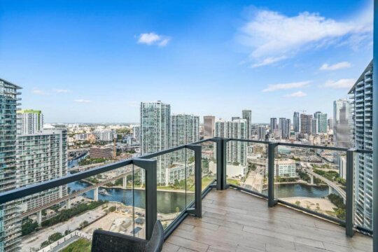 Panoramic view from a high-rise balcony in Miami, Florida, overlooking a cityscape with skyscrapers, a river, and a bridge under a bright blue sky with scattered clouds.