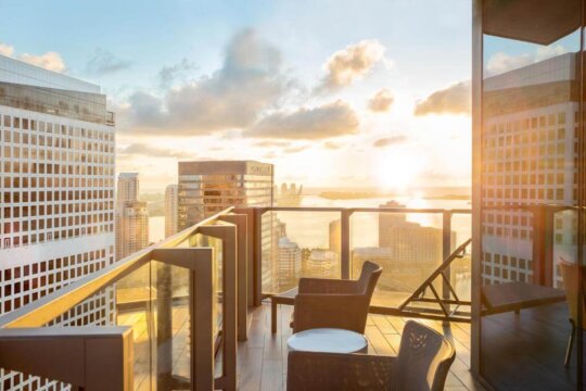 Sunrise over a cityscape viewed from a balcony with modern seating and glass railings, highlighting luxury high-rise buildings and a tranquil waterfront.