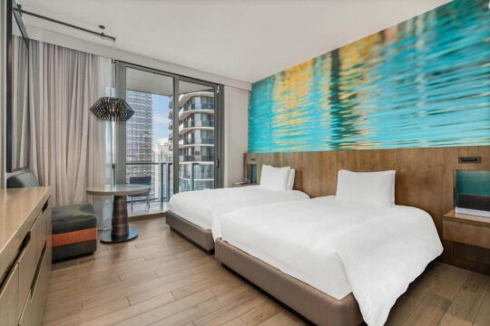 Modern hotel room with two queen beds, a vibrant abstract art mural of water, and a balcony with city views.