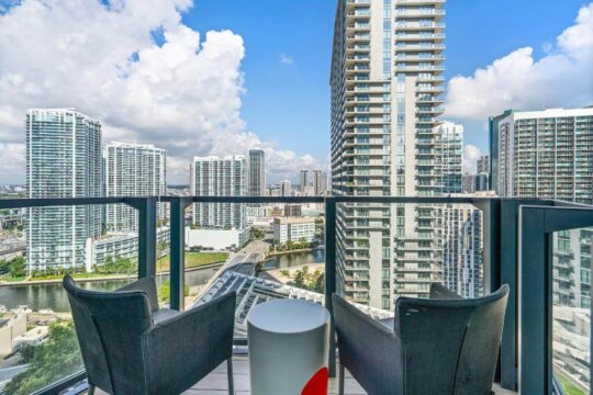Luxury balcony with two chairs and a small table overlooking a modern city skyline with tall skyscrapers and a river.