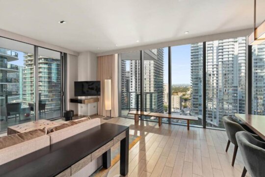Modern living room with floor-to-ceiling windows showcasing a panoramic view of a city skyline. A beige sectional sofa, sleek dark wood coffee table, and minimalist floor lamp create a stylish urban retreat.