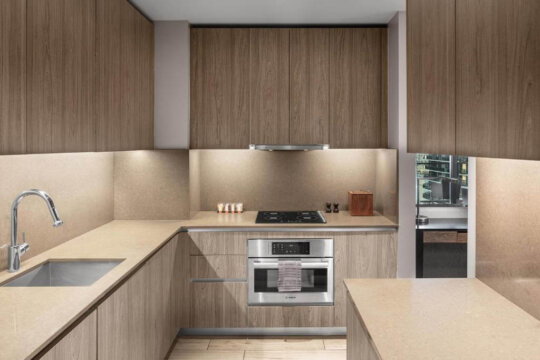 Modern kitchen with sleek, light wood cabinetry, a stainless steel oven, and a built-in stovetop. The countertop is a light, neutral stone, and under-cabinet lighting provides a warm glow.