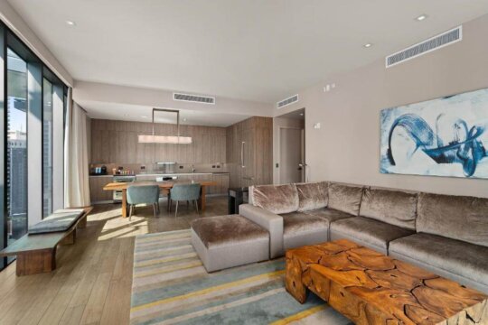 Modern open-plan living room and kitchen with a large sectional sofa, a rustic wooden coffee table, and a dining area with blue chairs, featuring large windows and contemporary art.