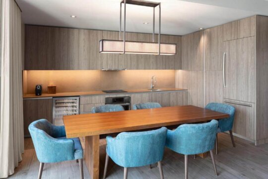 Modern dining room with a warm wood table and teal upholstered chairs, featuring sleek cabinetry, a wine fridge, and ambient lighting.