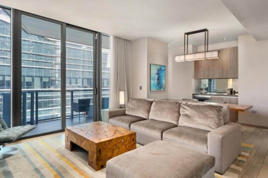 Modern living room with plush grey sectional sofa, unique carved wooden coffee table, and floor-to-ceiling windows overlooking a city skyline. Neutral color palette with pops of color in the abstract art and striped rug.