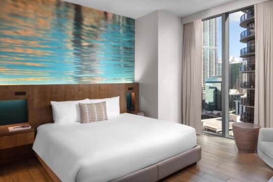 Modern hotel room with a king-size bed, vibrant abstract art on the wall, and a city skyline view through a large window.