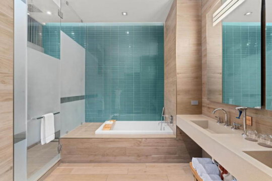 Luxurious modern bathroom with a deep soaking tub, teal subway tile accent wall, and a sleek concrete vanity with dual sinks. Warm wood tones and frosted glass shower enclosure add to the spa-like ambiance.