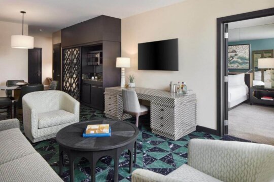A stylish hotel suite living area with a patterned desk, comfortable armchairs, a round coffee table, and a view into a bedroom.
