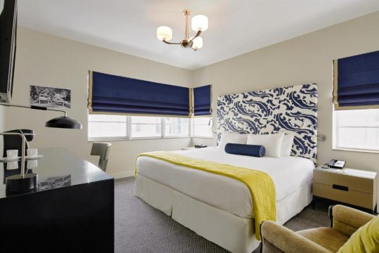 A modern hotel bedroom with a king-size bed featuring a blue and white patterned headboard, a yellow throw blanket, and a navy bolster pillow. Blue Roman blinds cover the windows, and a black desk with a lamp is visible to the left.