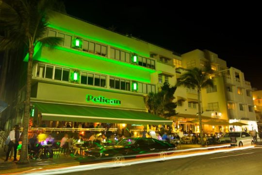 Nighttime view of a vibrant green illuminated building with a "Pelican" sign, outdoor seating, and palm trees, with cars and streetlights creating light trails on the road.
