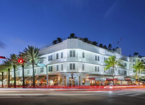 Bentley Hotel on Ocean Drive, Miami Beach, illuminated at dusk with palm trees and car light trails on the street.