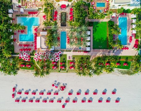 Aerial view of a luxury resort with vibrant red umbrellas, blue swimming pools, and lush green lawns leading to a sandy beach with rows of red lounge chairs.