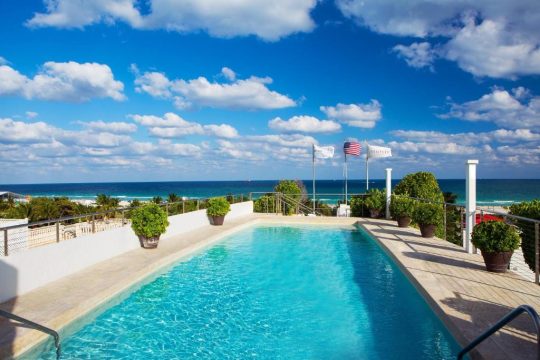 Rooftop swimming pool overlooking the ocean with lush green plants and flags, including the American flag and a flag with "BENTLEY" on it.