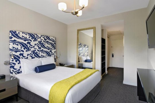 A modern hotel room features a king-size bed with a bold blue and white patterned headboard, a yellow throw blanket, and a dark blue bolster pillow. A large mirror reflects the room, showing a glimpse of a closet and a television.