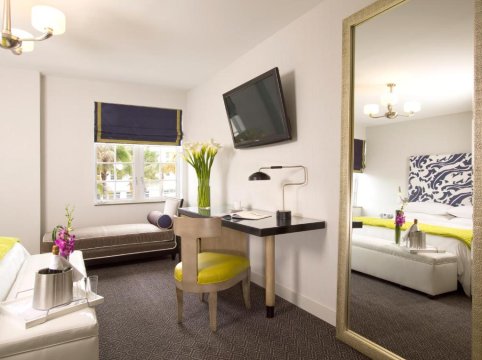 A modern hotel room with a desk, chair, and a large mirror reflecting a bed with a white and blue patterned headboard.