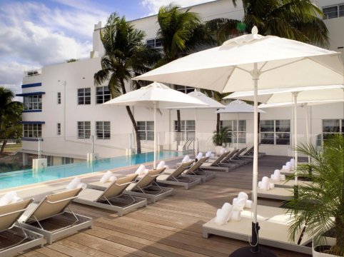 A sunny resort pool deck with white lounge chairs and umbrellas, overlooking a bright blue swimming pool and a white hotel building with palm trees.