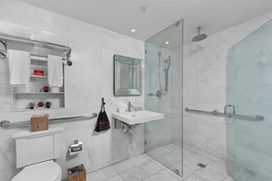 A modern, accessible hotel bathroom features a spacious walk-in shower with a glass enclosure and grab bars, a white toilet, and a sleek vanity with a large mirror, all finished with elegant marble tiling.