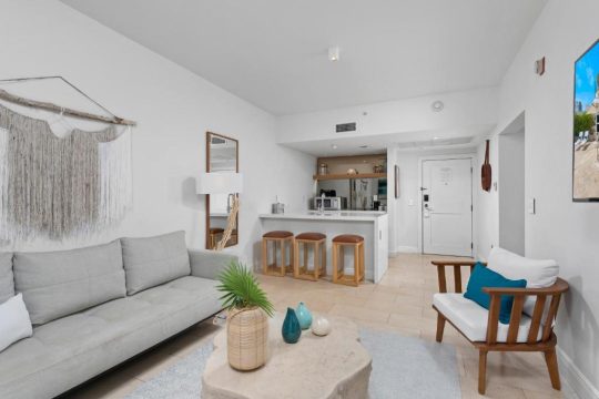Bright, modern living room with a grey sofa, woven wall art, and a minimalist kitchen bar with stools. Coastal decor elements like blue vases and a palm frond add to the relaxed vibe.