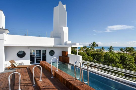 A modern white building with a rooftop pool and wooden deck overlooks a tropical beach with palm trees and the ocean under a clear blue sky.