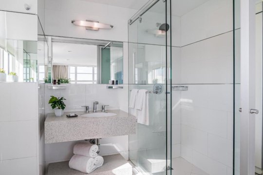 Modern bathroom with a sleek glass-enclosed shower, a granite-top vanity with a white sink, and neatly folded white towels. Natural light streams in, highlighting the clean, minimalist design.