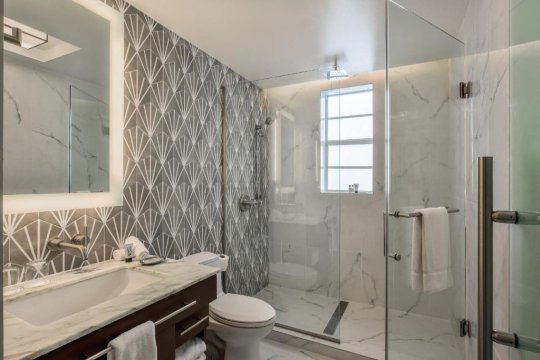 Modern bathroom with a sleek vanity, geometric patterned wallpaper, and a spacious glass-enclosed shower featuring marble-look tiles and a rainfall showerhead.