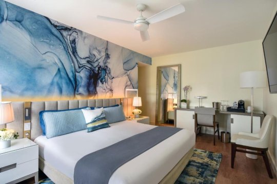 Modern hotel bedroom with a king-size bed, blue abstract art mural, and a well-lit workspace with a desk and chair.