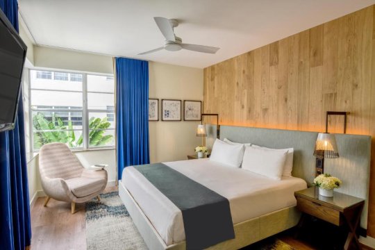 A modern hotel room featuring a king-size bed with white linens and a grey duvet, a plush armchair, and large windows with blue curtains, showcasing a bright and airy ambiance.