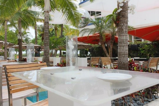 A modern outdoor dining area with a glass-top table set with white plates, surrounded by lush palm trees, tropical plants, and bright orange umbrellas, suggesting a vibrant vacation or resort setting.