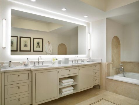 Luxurious hotel bathroom with a large, backlit mirror over a double vanity, featuring cream cabinetry, marble countertops, and a soaking tub with travertine tile.