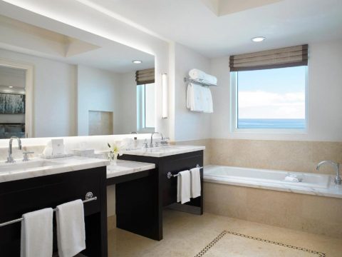 Luxurious hotel bathroom with dual vanities, a deep soaking tub, and a large window overlooking a serene ocean view.