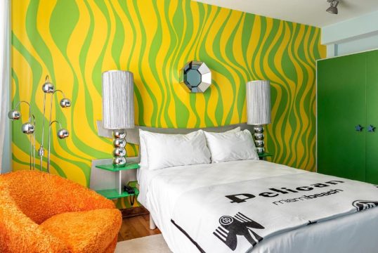 Retro bedroom with a vibrant yellow and green wavy wallpaper, orange shag armchair, and a bed with "Pelican Miami Beach" text on the duvet.